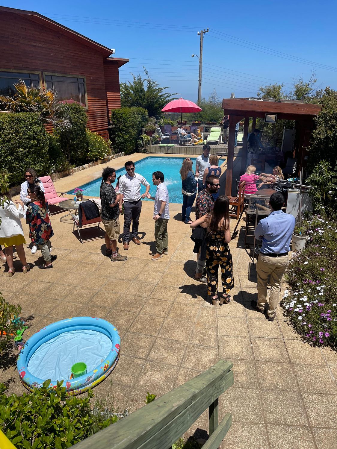 Piscina del hotel rodeada de jardín, con la casa de madera al fondo y huéspedes en la terraza